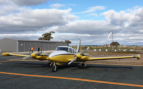 A yellow light aircraft sits on an airport tarmac with a tin hangar in the background